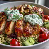 Overhead view of a vibrant Greek Chicken Power Bowl filled with quinoa, grilled chicken, crisp vegetables, Kalamata olives, fresh parsley, and a generous swirl of tzatziki sauce.