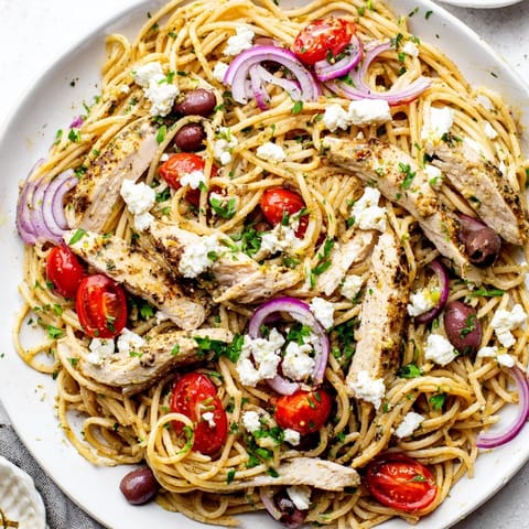 A steaming plate of Greek Chicken Spaghetti features ripe cherry tomatoes, crisp red onion, fresh parsley, and a bright lemon-oregano dressing.  