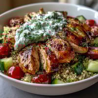 Close-up of the Greek Chicken Power Bowl featuring golden, juicy lemon-herb chicken slices, fluffy quinoa, diced cucumbers, halved cherry tomatoes, and a dollop of creamy tzatziki.